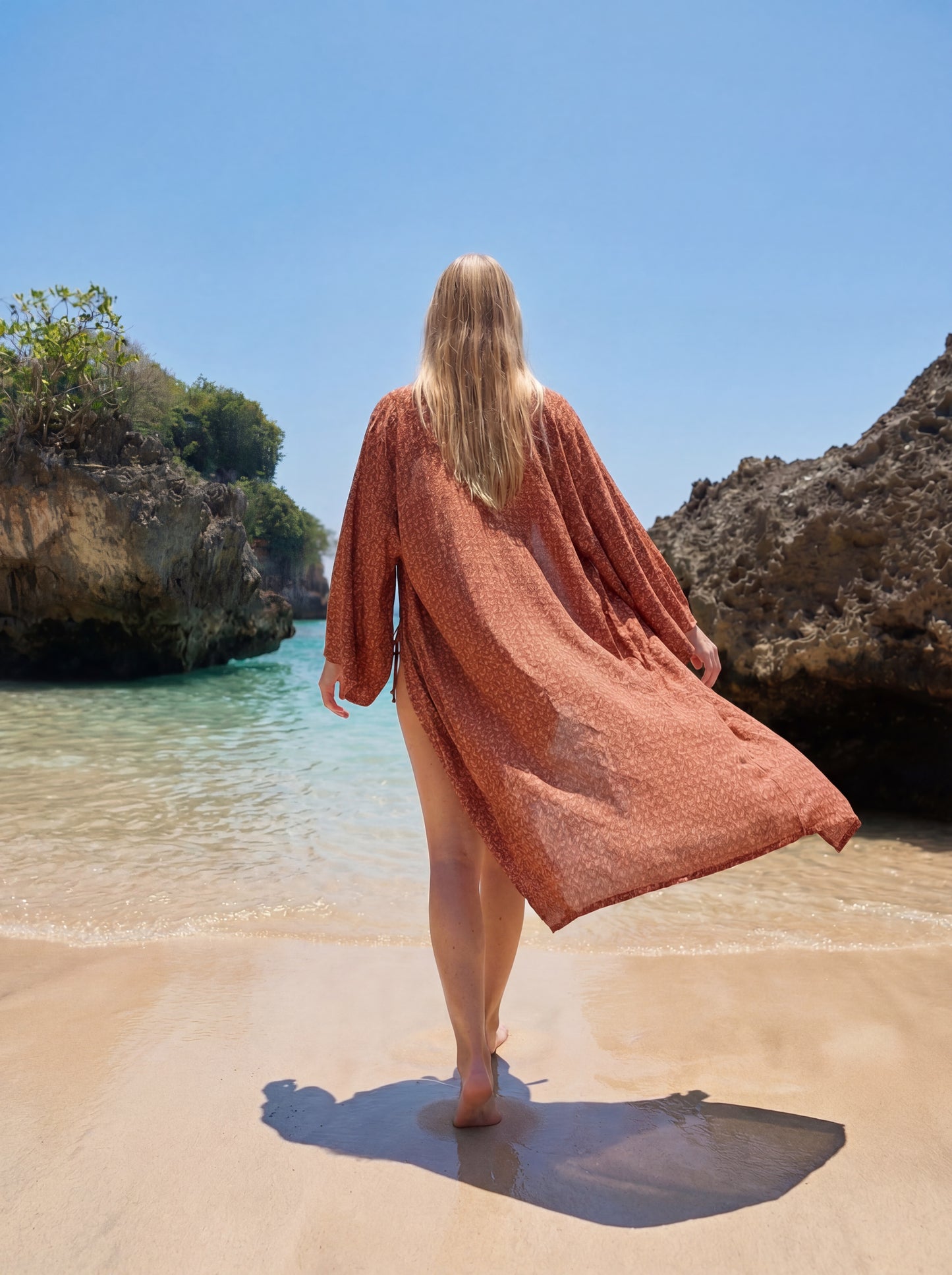Woman in a rust-colored cover-up walking on a sandy beach with clear blue water and rocky cliffs.