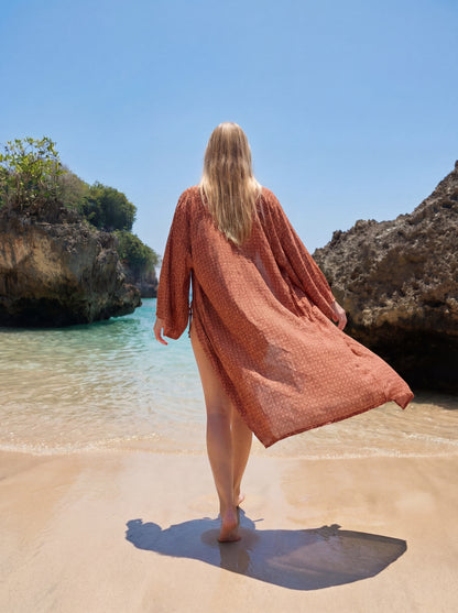 Woman in a rust-colored cover-up walking on a sandy beach with clear blue water and rocky cliffs.